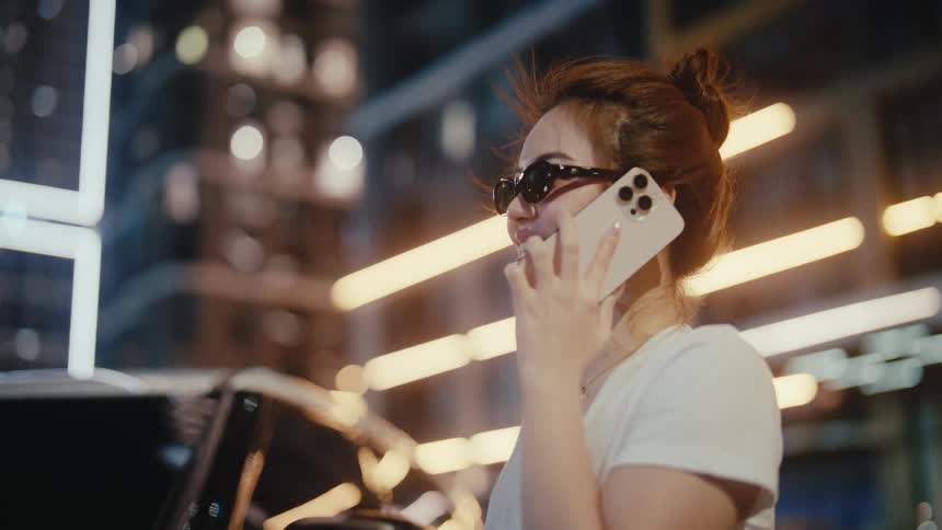 A stylish Asian woman in sunglasses smiles while talking on her smartphone at night, with the city's vibrant lights creating a beautiful bokeh background