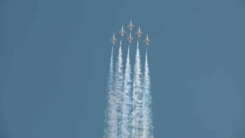A squadron of six fighter jets in perfect formation performs a vertical climb, leaving impressive white smoke trails against the vast blue sky