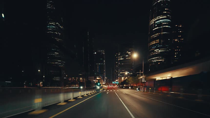 A point-of-view shot from a car driving through the glowing streets of a futuristic city at night surrounded by towering skyscrapers