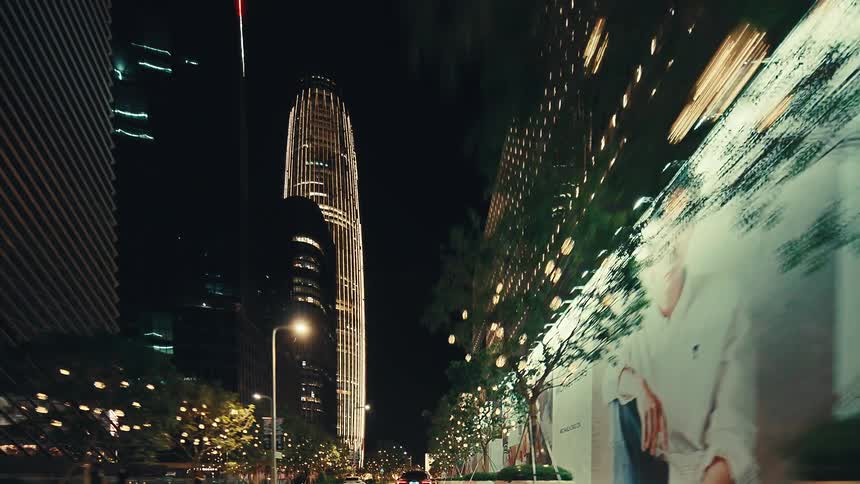 A smooth forward tracking shot through a modern city street at night, with towering skyscrapers and trees adorned with lights