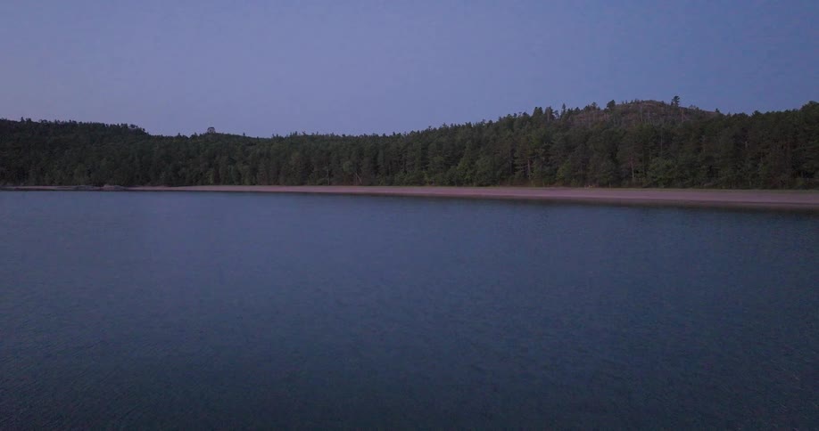 A tranquil aerial flight over a serene lake towards a sandy beach and lush forest at twilight
