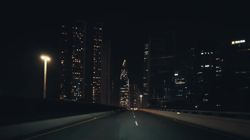 Point-of-view of a car driving along a highway at night, approaching the brightly illuminated, futuristic skyline of a modern city