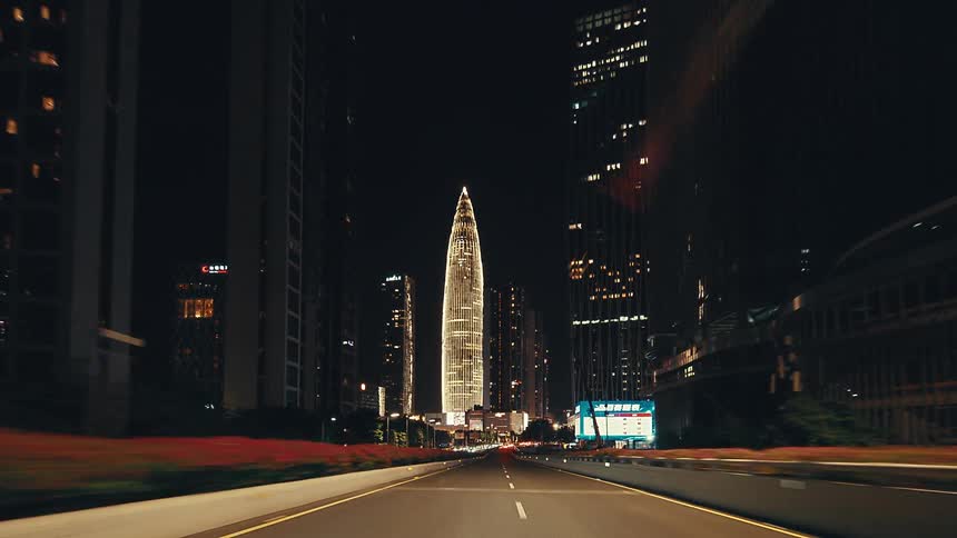 Point-of-view of a car driving along a highway at night, approaching the brightly illuminated, futuristic skyline of a modern city