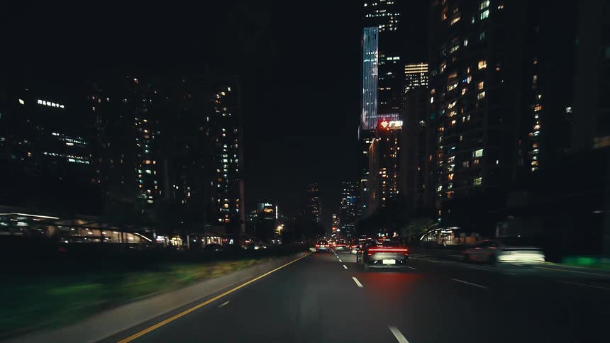 Driving through the brightly lit streets of a modern metropolis at night, with towering skyscrapers and steady traffic creating a dynamic urban scene