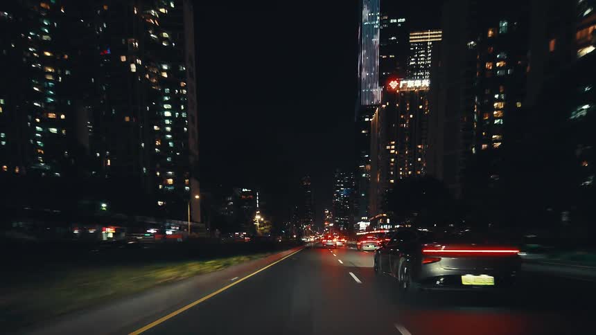 Driving through the brightly lit streets of a modern metropolis at night, with towering skyscrapers and steady traffic creating a dynamic urban scene