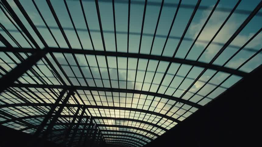 A smooth tracking shot looks up at the sky and clouds through the silhouetted framework of a modern curved glass roof