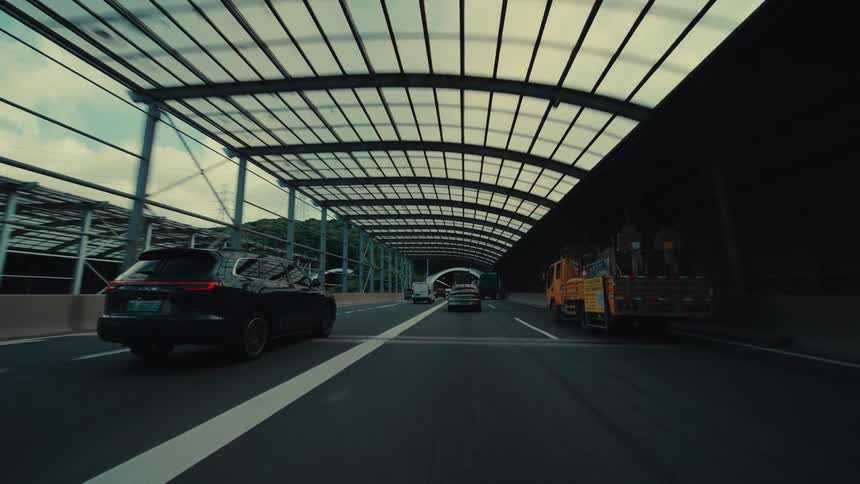 A cinematic point-of-view shot from a car driving on a modern highway, passing under a translucent roof before heading towards a tunnel