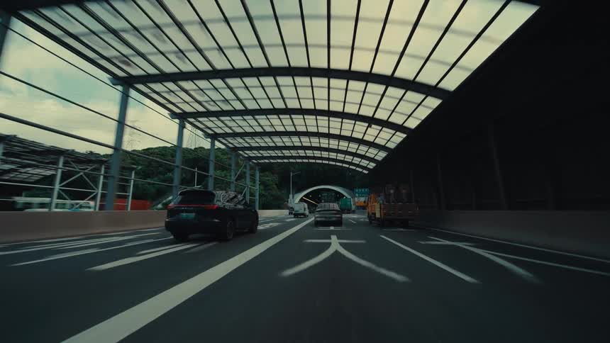 A cinematic point-of-view shot from a car driving on a modern highway, passing under a translucent roof before heading towards a tunnel