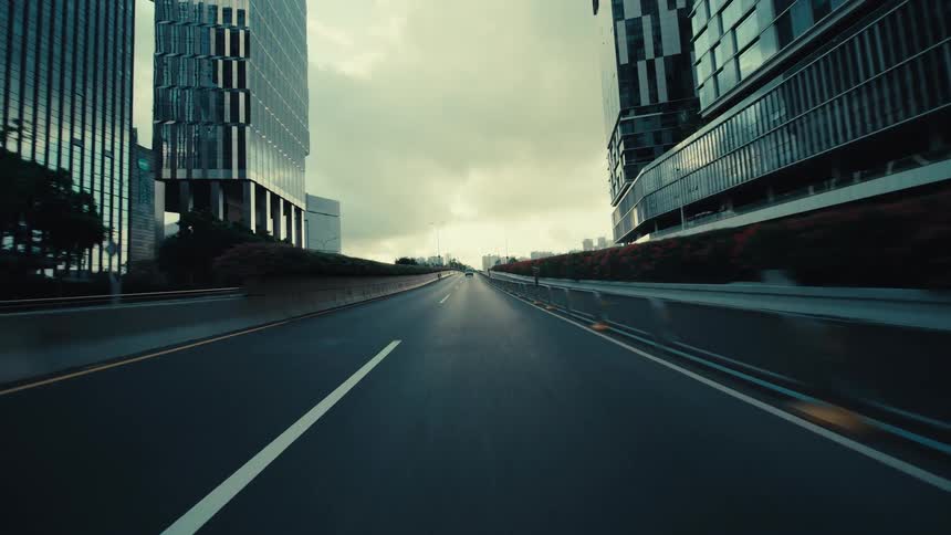 A low-angle POV shot driving down an empty highway through a modern city of glass skyscrapers on a cloudy day