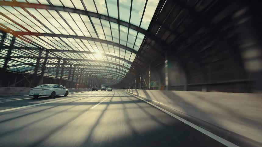 A fast-paced, low-angle point-of-view drive through a modern highway with a translucent arched canopy as sunlight creates dynamic shadows and lens flare