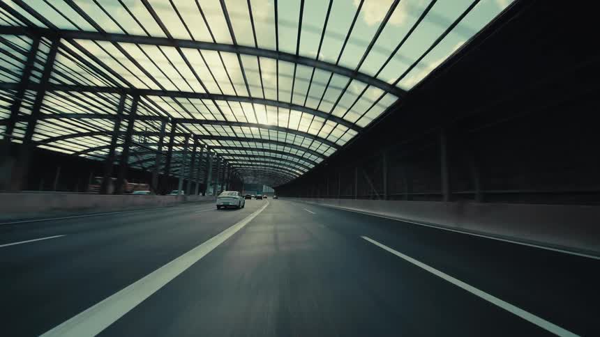 A fast-paced, low-angle point-of-view drive through a modern highway with a translucent arched canopy as sunlight creates dynamic shadows and lens flare