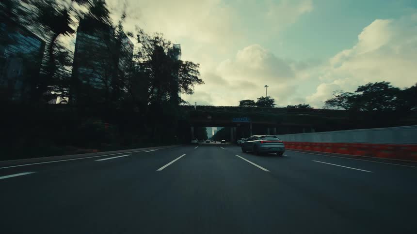 A cinematic point-of-view drive on a wide city highway approaching a modern skyline through lush green trees and under an overpass