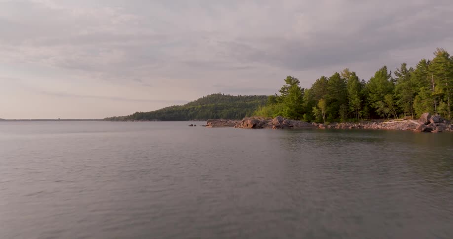 An aerial view flies over a calm lake towards a rugged, tree-lined coastline during a peaceful sunset