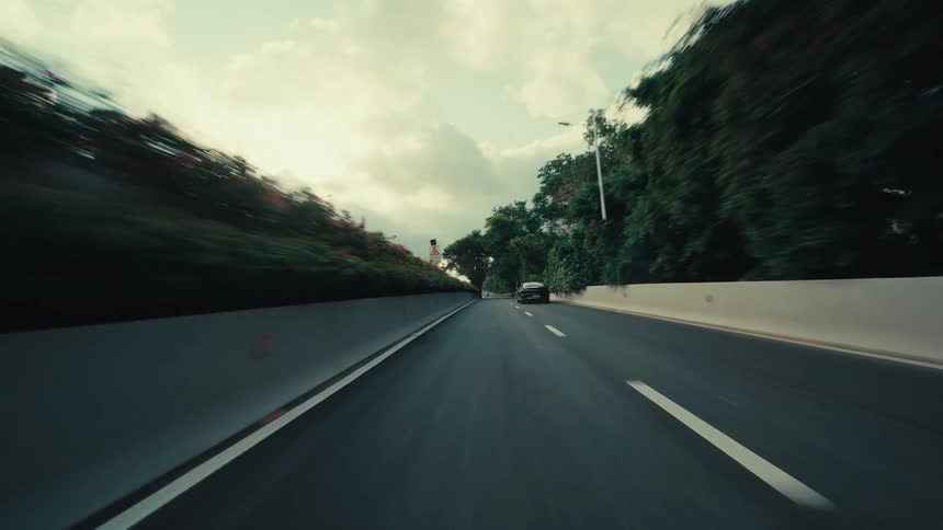 A cinematic point-of-view shot of a car driving fast out from under an overpass onto a modern highway surrounded by lush greenery