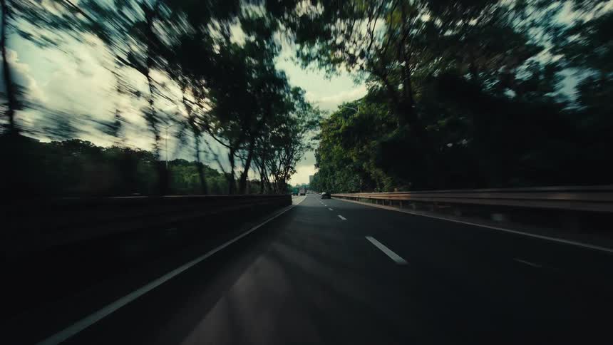 A cinematic point-of-view shot of a car driving fast out from under an overpass onto a modern highway surrounded by lush greenery