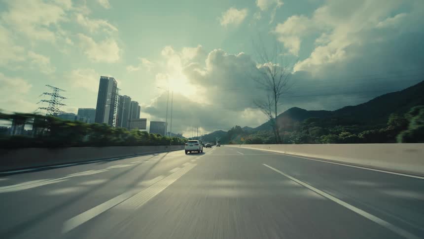 Cinematic low-angle POV shot driving on a highway towards a modern city with skyscrapers and mountains under a cloudy sky