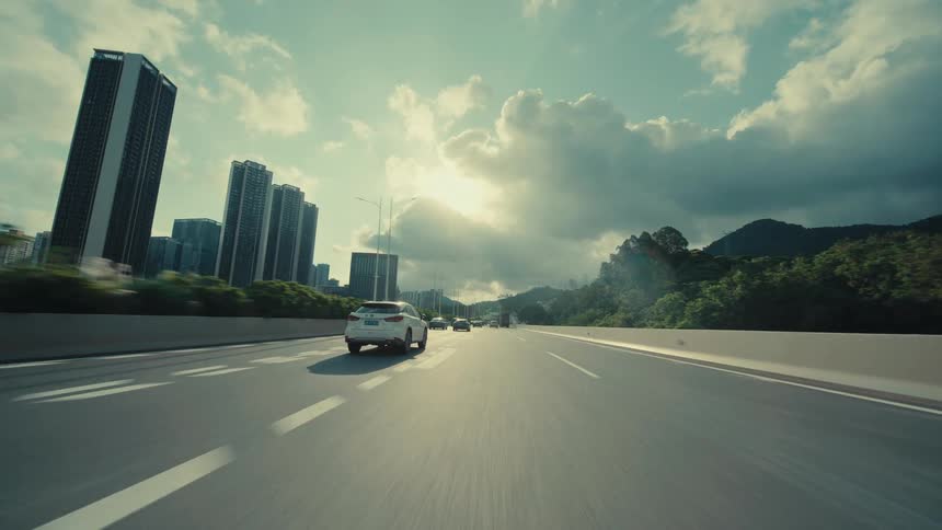 Cinematic low-angle POV shot driving on a highway towards a modern city with skyscrapers and mountains under a cloudy sky