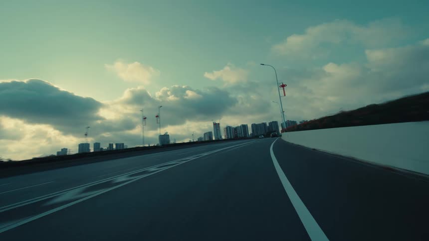 A moody, cinematic point-of-view of a car driving along a quiet highway, emerging from under an overpass to reveal a distant modern city skyline