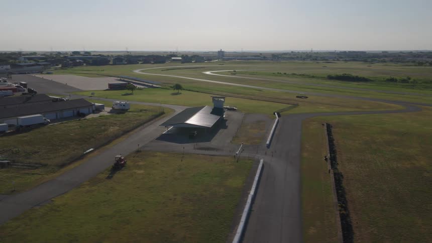 A slow, cinematic drone shot orbits an empty motorsports race track with a control tower on a sunny day
