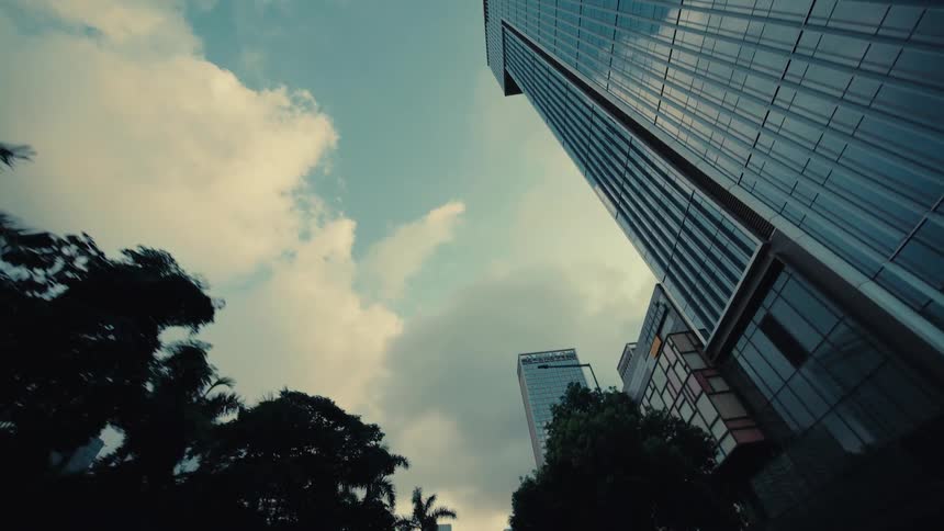 A stylized, low-angle tracking shot moves backward past silhouetted palm trees, revealing a modern glass skyscraper against a cloudy sky