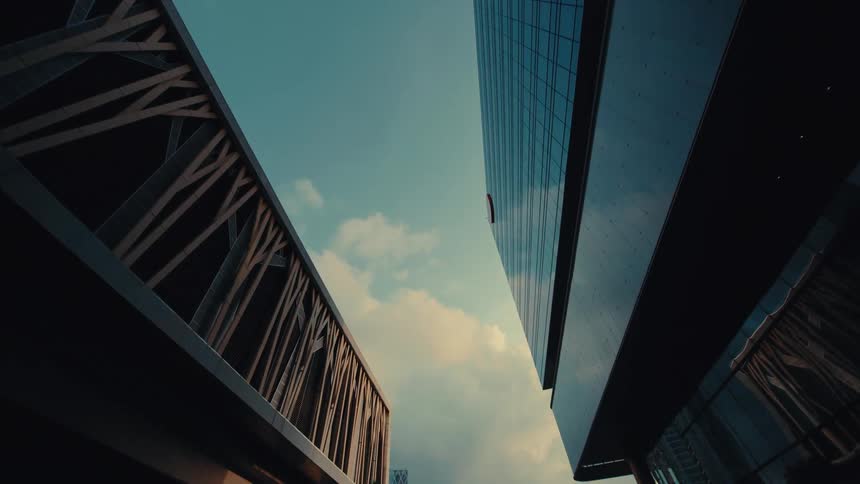 A low-angle dolly shot pulls back to reveal two modern skyscrapers with a walkway between them against a moody sky