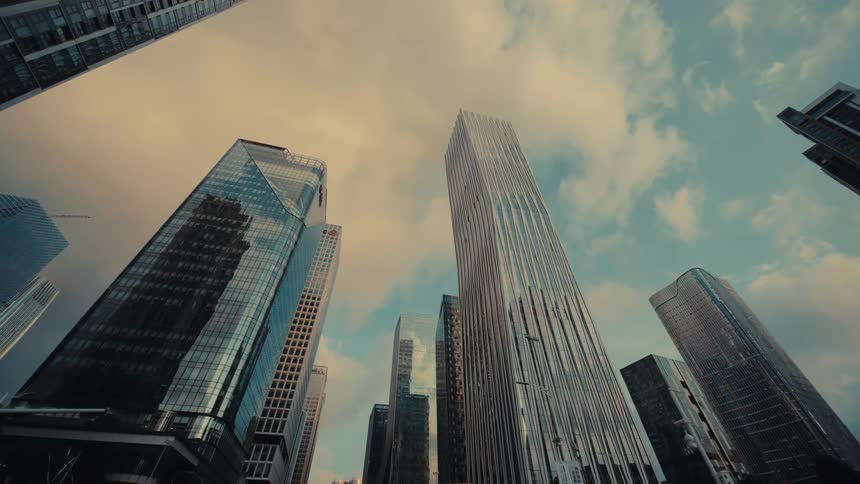 A cinematic low-angle dolly shot glides forward through a modern city, looking up at impressive glass skyscrapers against a cloudy sky
