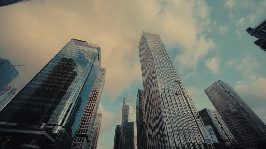 A cinematic low-angle dolly shot glides forward through a modern city, looking up at impressive glass skyscrapers against a cloudy sky