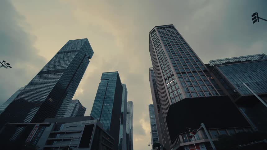 Driving through a modern downtown metropolis while looking up at imposing skyscrapers under a gloomy, overcast sky