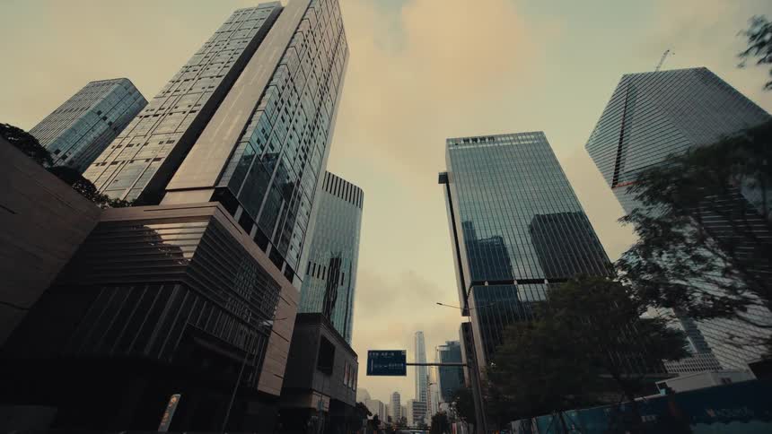 A low-angle dolly shot looking up at modern glass skyscrapers lining a city street at sunset