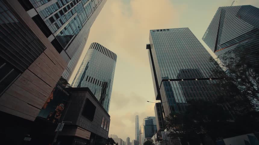 A low-angle dolly shot looking up at modern glass skyscrapers lining a city street at sunset