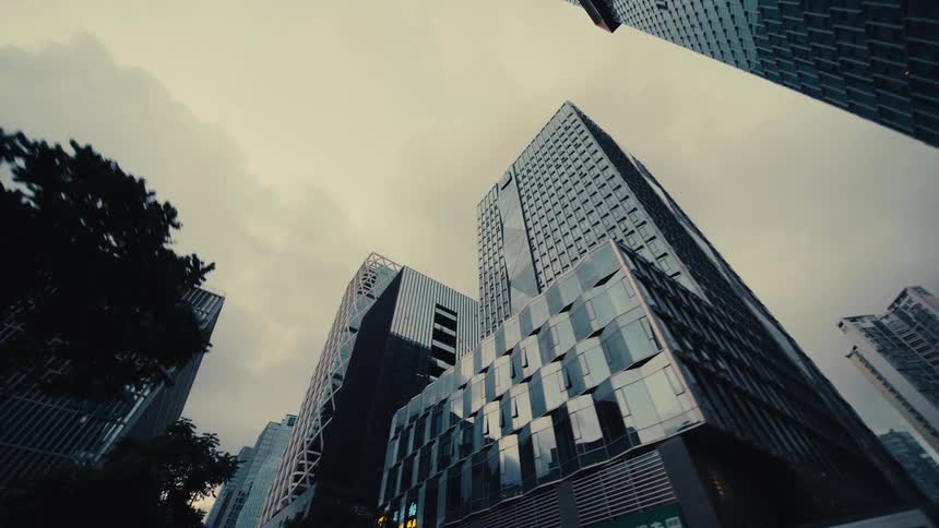 A low-angle tracking shot glides through a modern city canyon of towering glass skyscrapers under an overcast sky