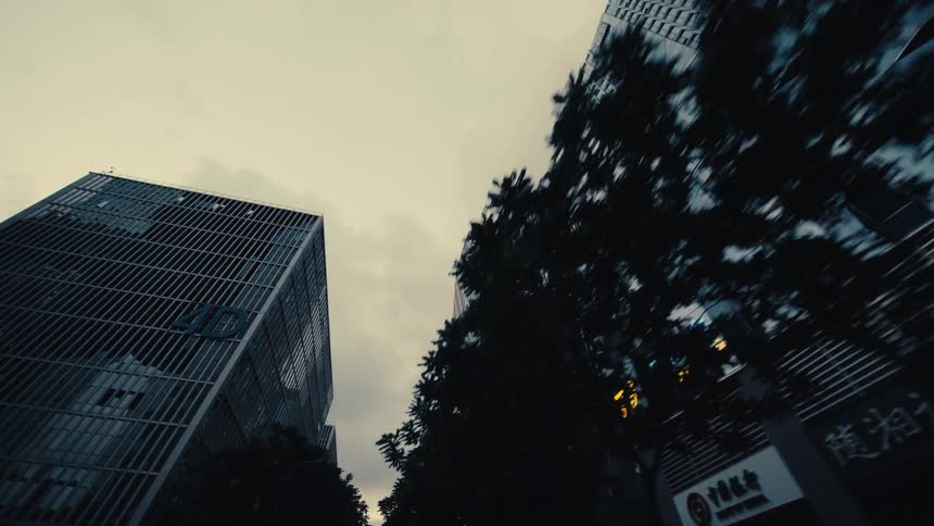 A low-angle tracking shot glides through a modern city canyon of towering glass skyscrapers under an overcast sky