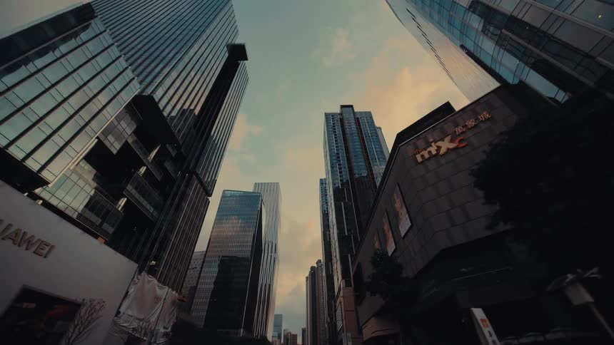 A low-angle reverse tracking shot looking up at the towering glass skyscrapers of a modern city's business district at dusk