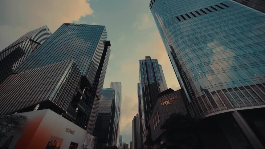 A low-angle reverse tracking shot looking up at the towering glass skyscrapers of a modern city's business district at dusk