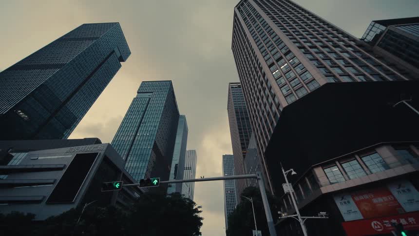 A slow, low-angle reverse dolly shot reveals towering modern skyscrapers reaching into a cloudy sky from a city street