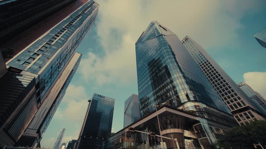 A low-angle tracking shot reveals the impressive modern glass skyscrapers of a city's financial district against a blue sky