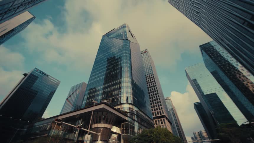 A low-angle tracking shot reveals the impressive modern glass skyscrapers of a city's financial district against a blue sky