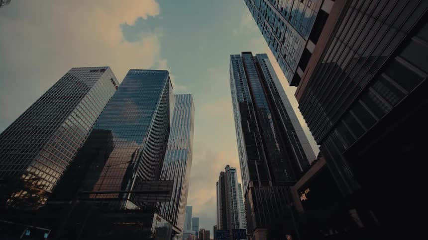A low angle moving shot looks up at towering modern skyscrapers against a warm sunset sky in a bustling city