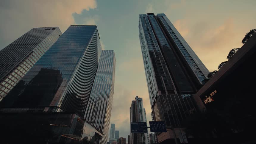 A low angle moving shot looks up at towering modern skyscrapers against a warm sunset sky in a bustling city