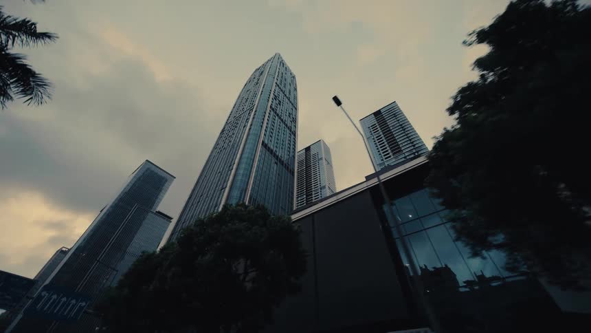 A dramatic, slow pull-back shot from a low angle reveals towering modern skyscrapers in a city under a cloudy sky