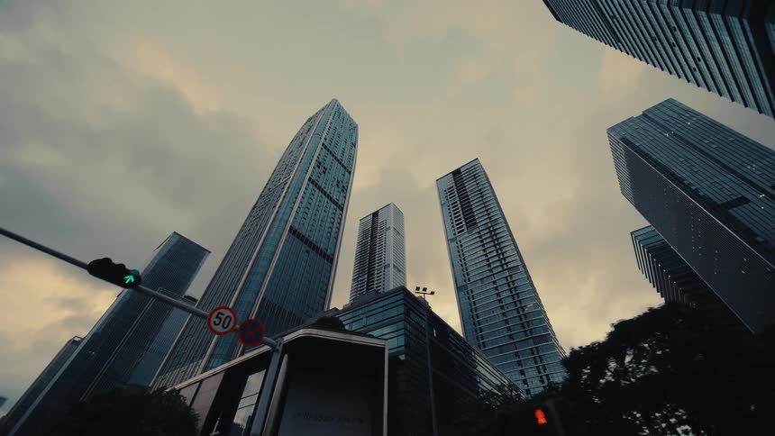 A dramatic, slow pull-back shot from a low angle reveals towering modern skyscrapers in a city under a cloudy sky