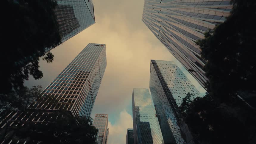 A slow-moving, low-angle driving shot looking up at a modern city skyline of glass skyscrapers against a cloudy, warm-toned sky
