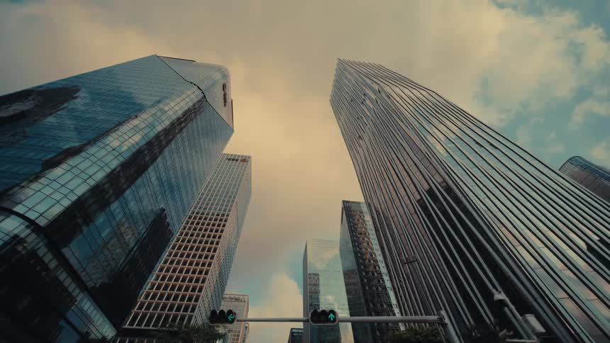 A slow-moving, low-angle driving shot looking up at a modern city skyline of glass skyscrapers against a cloudy, warm-toned sky
