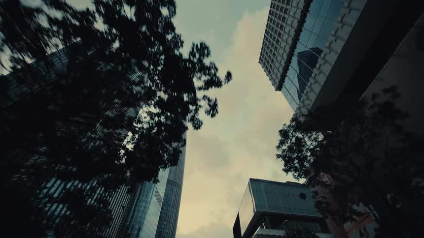 A low-angle reverse dolly shot glides past towering glass skyscrapers in a futuristic city at dusk