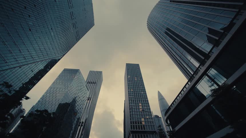 A low-angle dolly shot reveals the towering and modern glass skyscrapers of a financial district against a gloomy, overcast sky