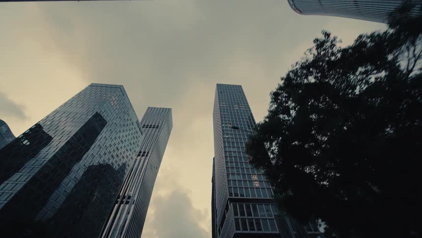 A low-angle dolly shot reveals the towering and modern glass skyscrapers of a financial district against a gloomy, overcast sky