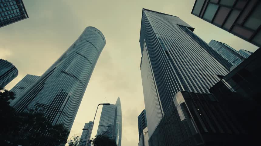 A low-angle forward tracking shot reveals the imposing modern skyscrapers of a city's business district under an overcast sky