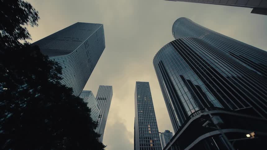 A slow dolly-out, low-angle shot reveals towering modern glass skyscrapers against a moody, overcast sky