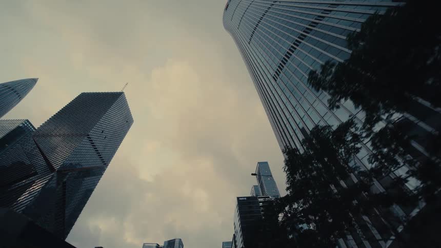 A low-angle driving shot through a modern city looks up at towering glass skyscrapers against an overcast sky