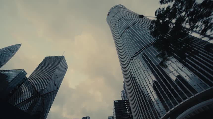 A low-angle driving shot through a modern city looks up at towering glass skyscrapers against an overcast sky